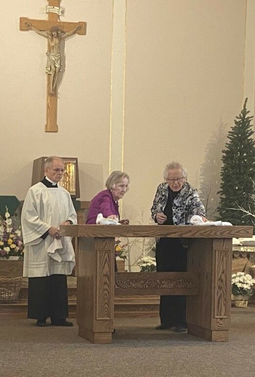Three people stand at a church altar beneath a crucifix, with candles and floral decorations nearby.