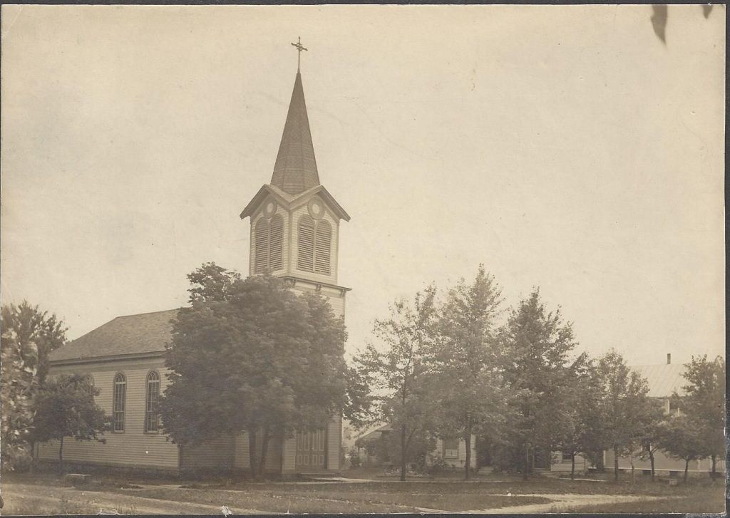 Old sepia photo of a church with a tall steeple among trees and a small building nearby