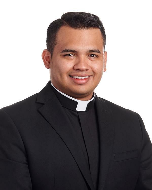 Portrait of a smiling clergyman in a black clerical shirt with white collar against a blue backdrop