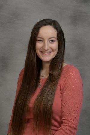 Smiling woman with long brown hair wearing a coral top against a gray backdrop