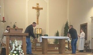 People arranging a church altar beneath a crucifix, with a priest at the lectern and candles nearby.