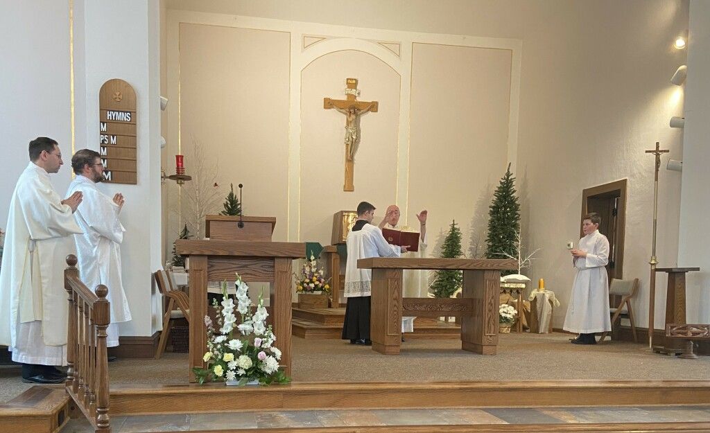 Church altar with crucifix, clergy in white robes, and a person seated at the lectern during a service