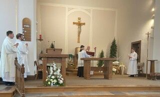 Church altar with crucifix; priest and altar servers stand during Mass beside white floral arrangements.