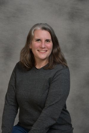 Smiling person in a dark gray sweater seated against a gray studio backdrop