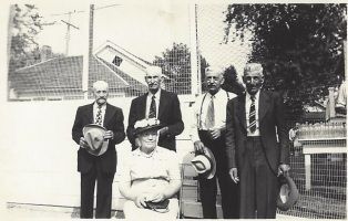Group of five people in formal attire posing outdoors, two seated in front and three standing behind them.