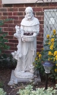 White statue of a robed man holding a basket in a garden beside a brick wall and flowers