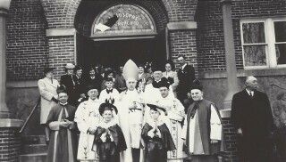Black-and-white group photo of people posed outside a brick building entrance, some in formal attire and a bishop’s hat