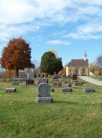 Gravestones in a grassy cemetery under a blue sky, with autumn trees and a small church in the background