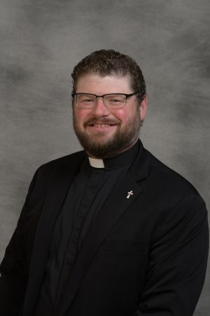 Portrait of a smiling person in black clerical attire against a gray studio backdrop