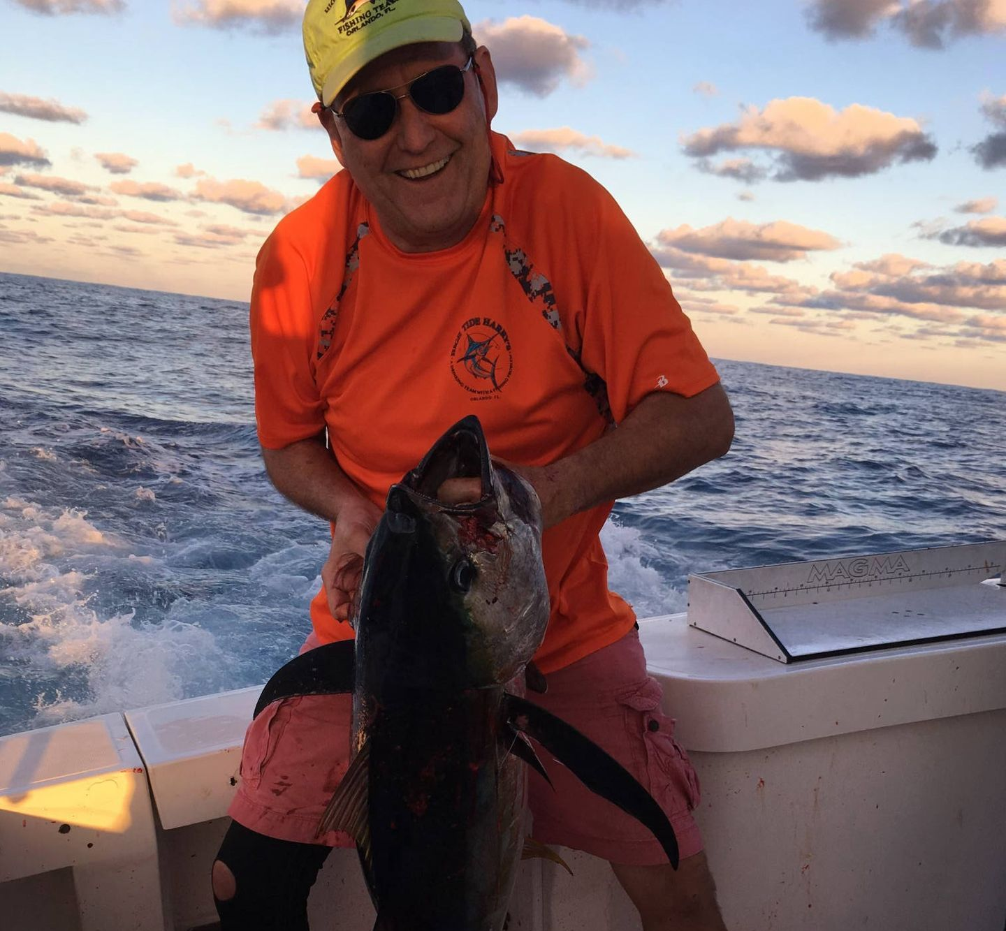 Harry from High tide Harry's on a boat holding a large fish; orange shirt, pink shorts, sunglasses, ocean background.