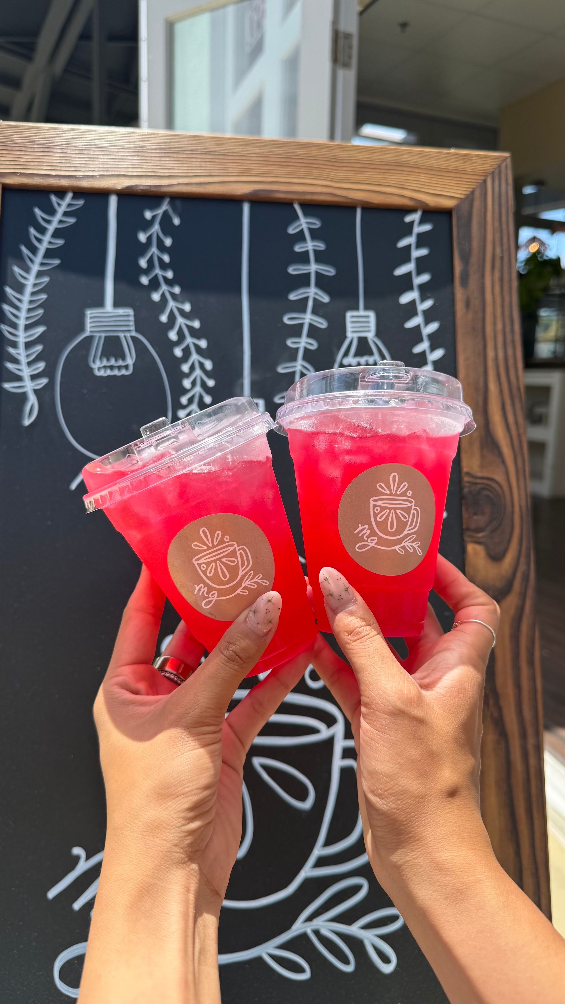 Two hands holding up red drinks in front of a chalkboard sign with floral drawings.