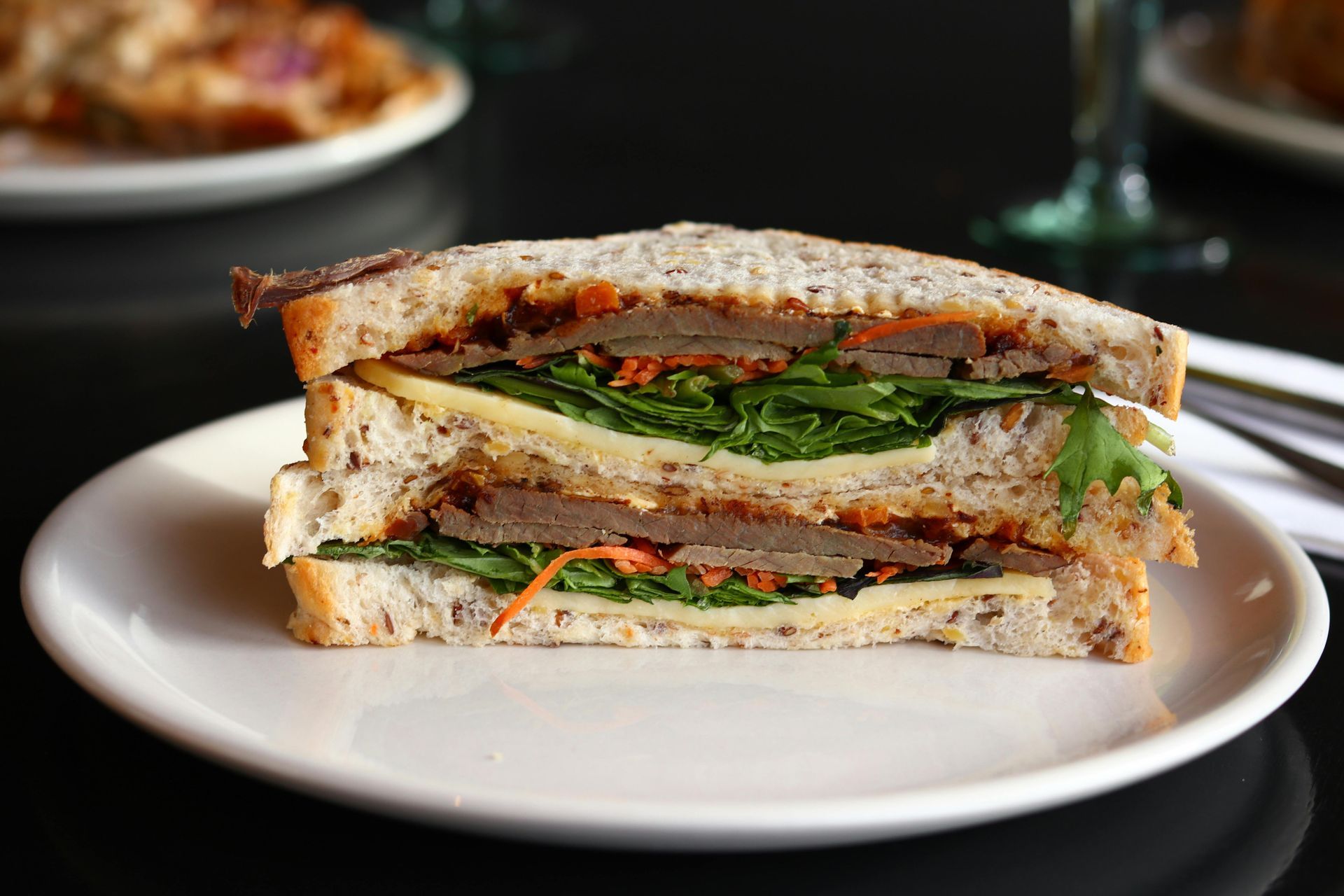 Sandwich on a white plate, cross-section view: bread, meat, cheese, greens, and carrots.