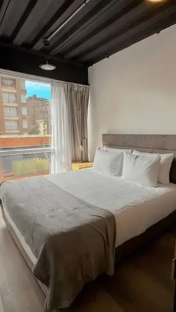 Apartment suite in Bogotá with a double bed, white linens, gray curtains, and a window with city views.