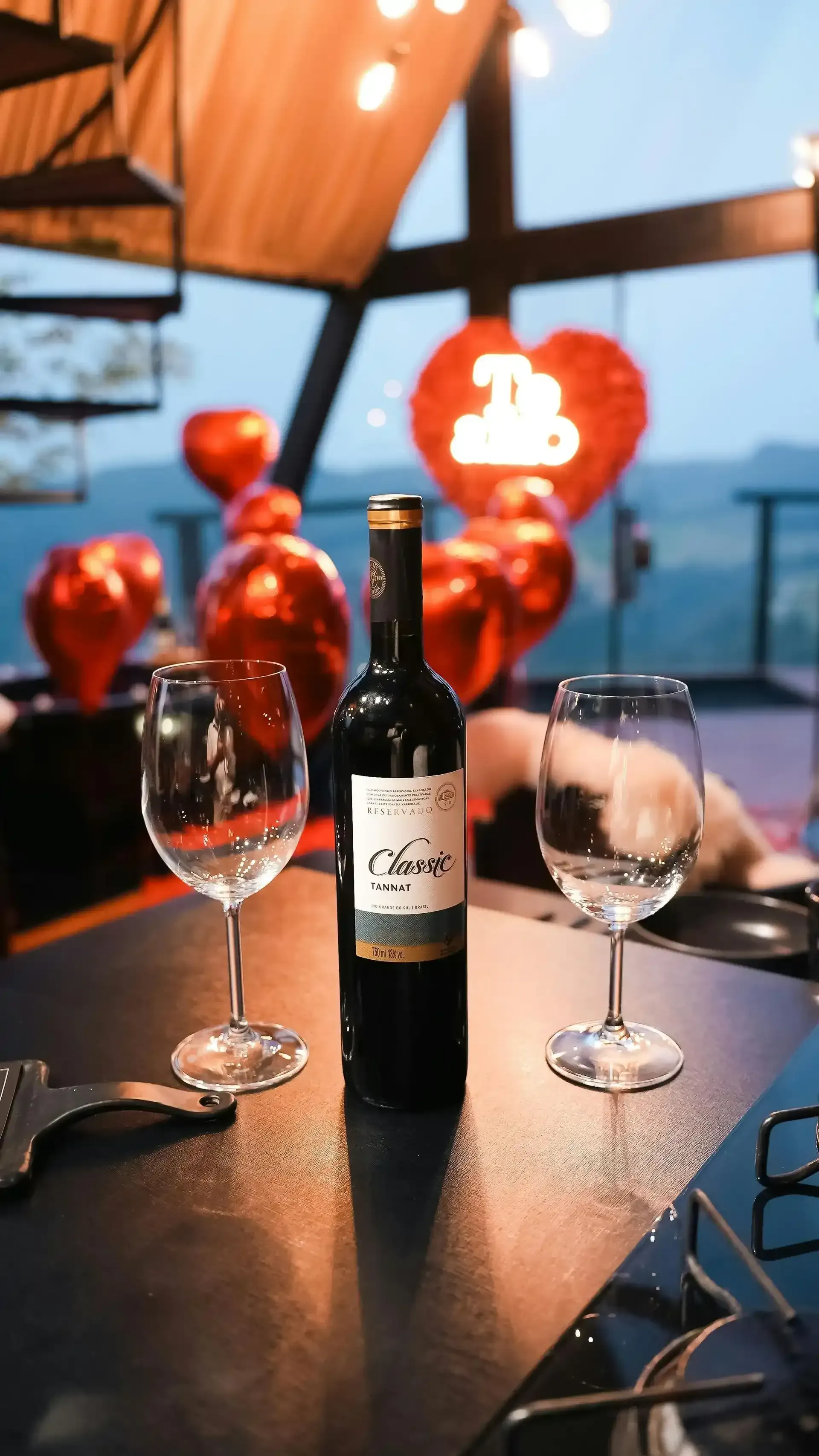 Botella de vino y dos copas sobre mesa con globos en forma de corazón, plan romántico en pareja en Bogotá.