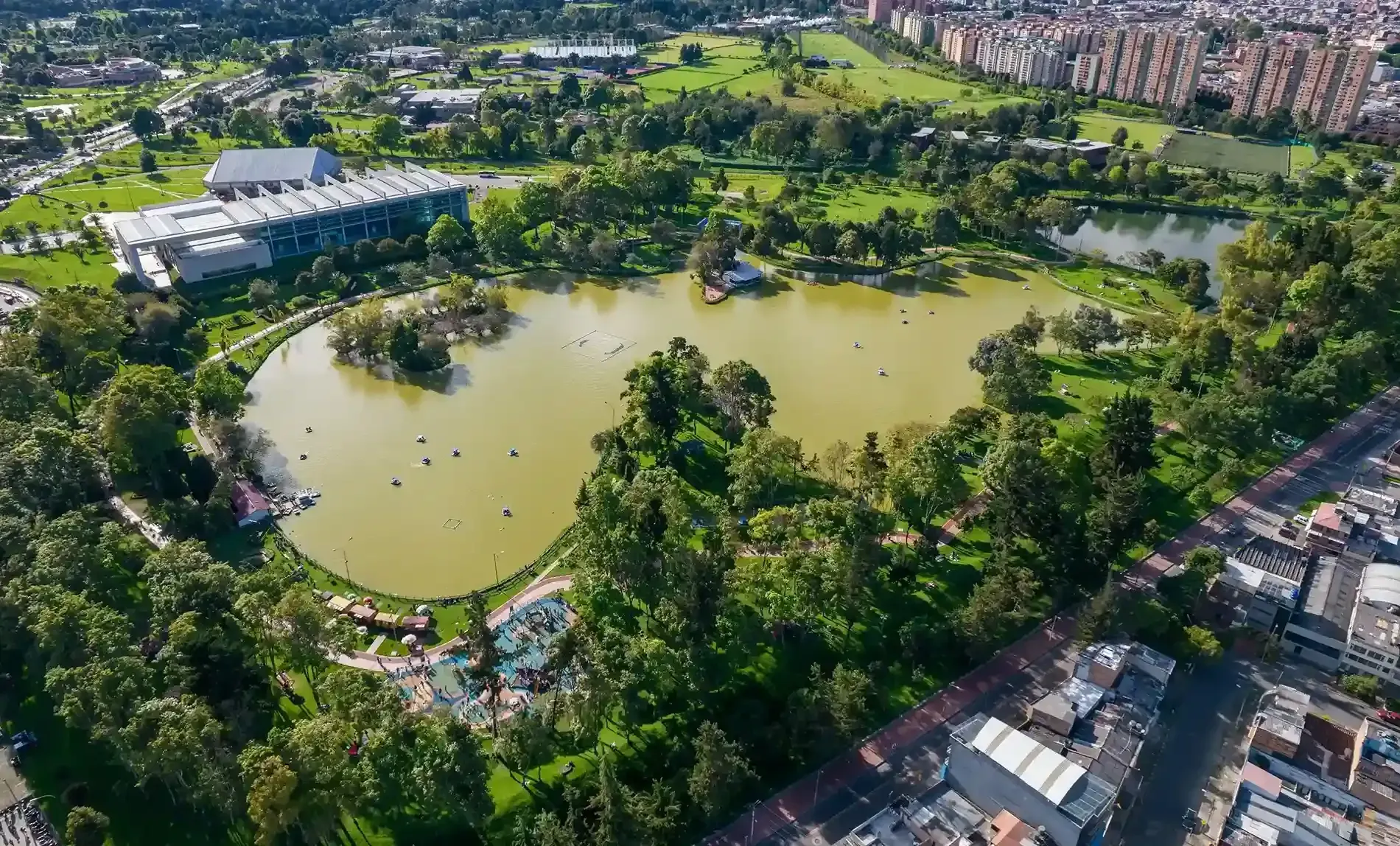 Vista aérea del Parque de los Novios en Bogotá, mostrando el gran lago rodeado de árboles y senderos, con edificios modernos y residenciales al fondo