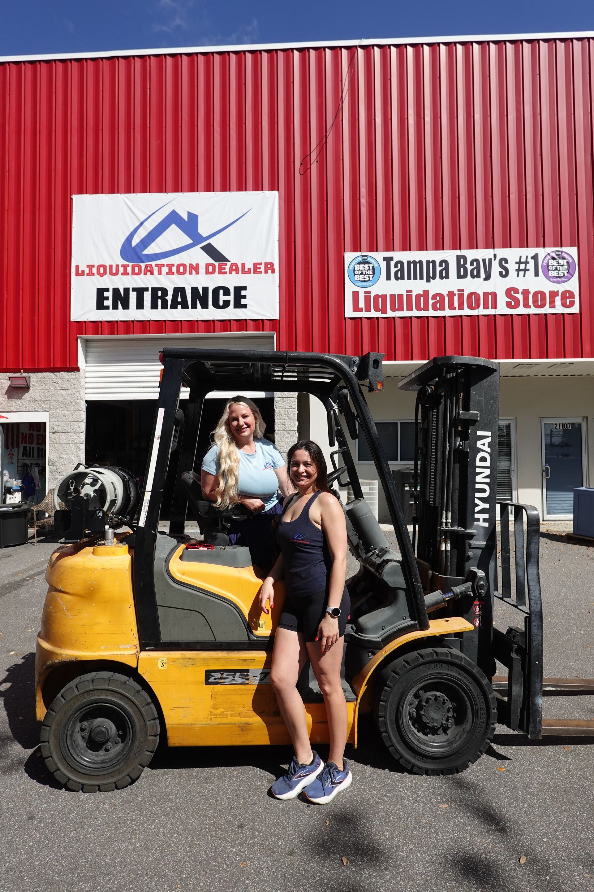 Two people by a yellow forklift outside a liquidation store entrance with red corrugated siding.