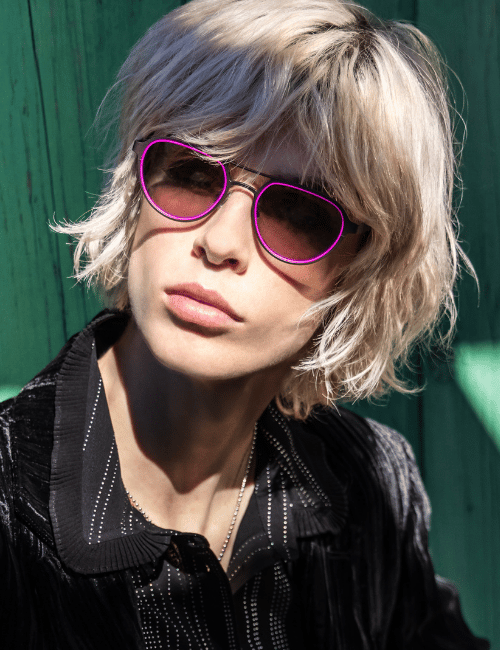 Person with blonde hair wearing purple sunglasses and a black shirt against a green background