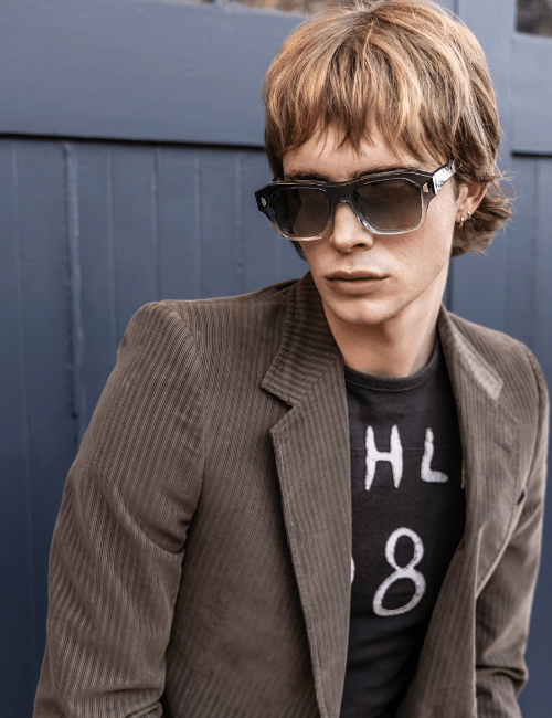 Person in sunglasses and brown blazer over a black shirt, posing against a gray wall.