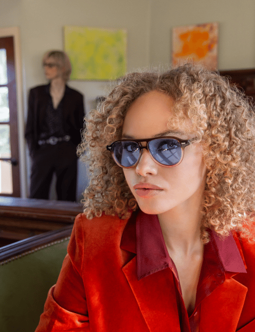Person in red blazer and blue sunglasses sitting indoors; blurred figure and colorful art in background