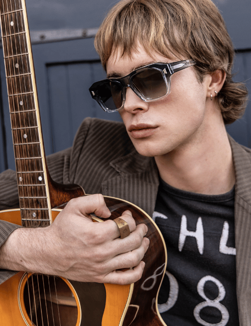 Person in sunglasses holding a guitar, wearing a dark shirt against a blue-gray background.