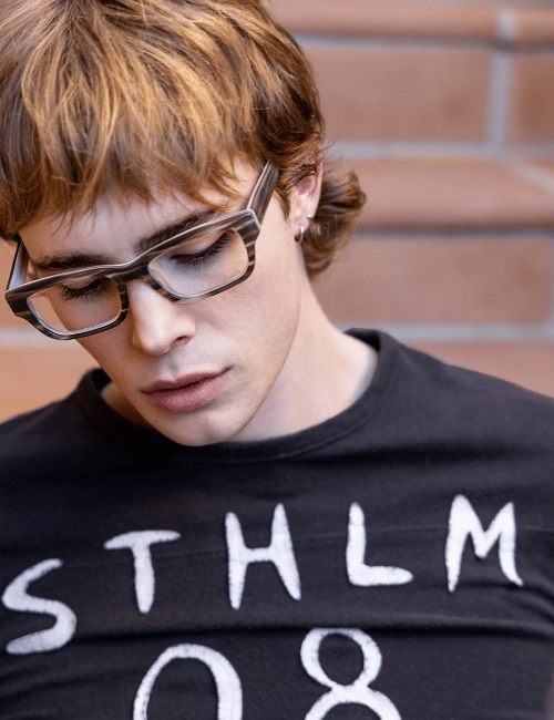 Person with glasses and black shirt reading “STHLM 08” against a brick wall.