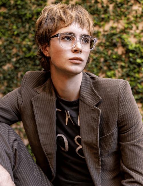 Person with short brown hair and glasses wearing a brown blazer, seated outdoors against a leafy background.