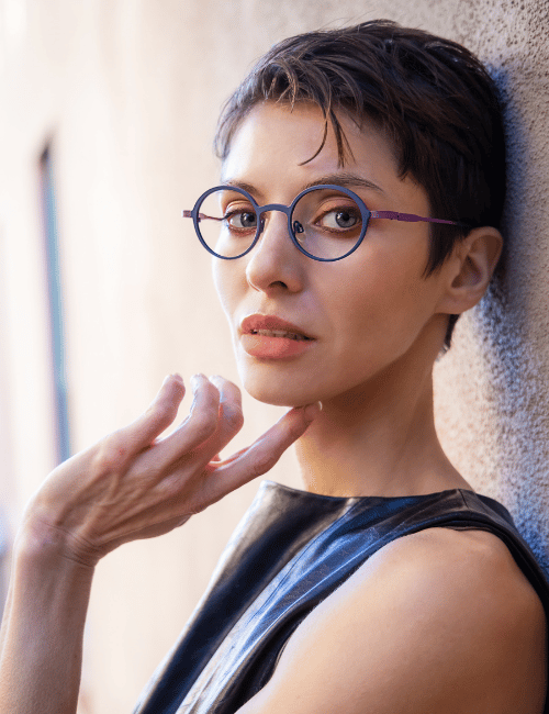 Person with short dark hair and round glasses, posing beside a textured wall in a sleeveless dark top.