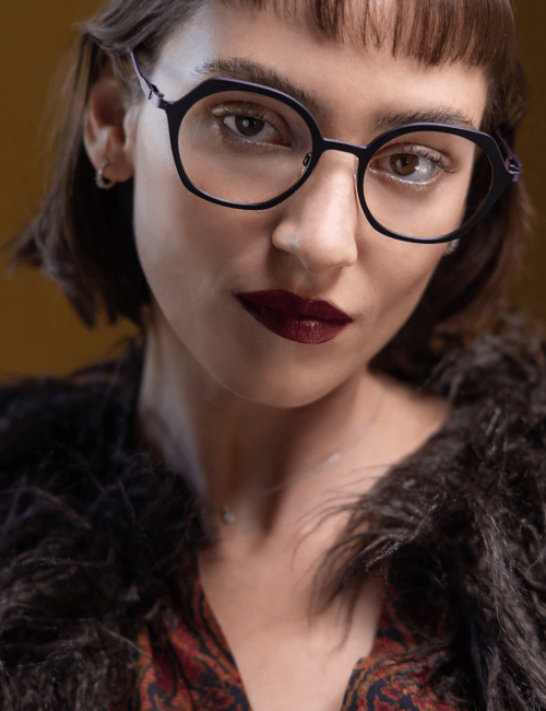 Close-up portrait of a person with round black glasses, short brown hair, and dark lipstick wearing a fur-trimmed coat.