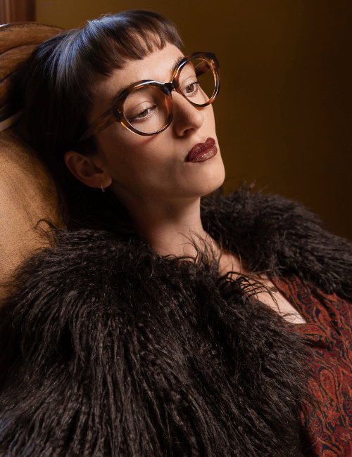 Portrait of a person with glasses and dark fur coat against a warm brown background