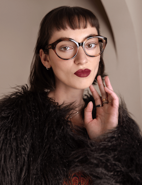 Portrait of a person with round glasses, dark bangs, red lipstick, and a black faux-fur coat against a beige backdrop