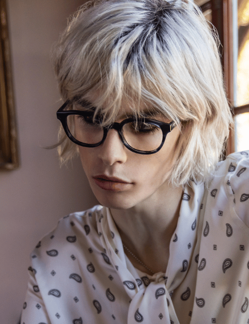 Person with black glasses and short blond hair, wearing a white patterned shirt, looking down by a window.