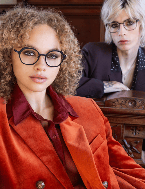 Two women in glasses pose indoors, one in a red jacket foreground and one seated behind in dark clothing.