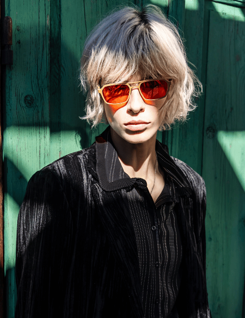 Person with orange sunglasses and tousled blond hair against a green wooden wall