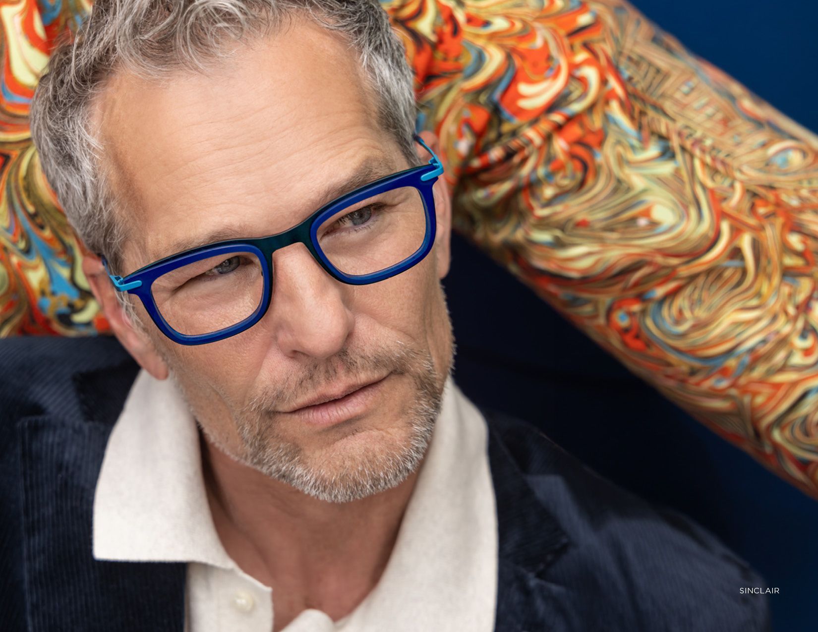 Man with blue glasses in a dark jacket, seated against a colorful patterned background