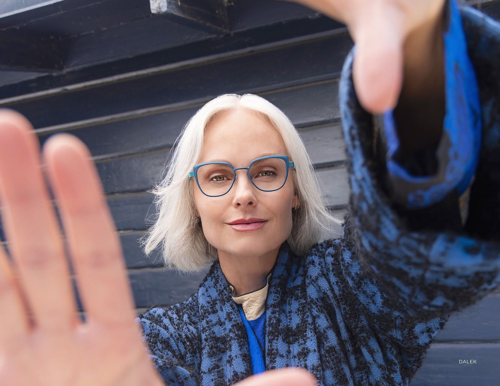 Woman with white hair and blue glasses reaching toward the camera, wearing a blue patterned jacket outdoors.