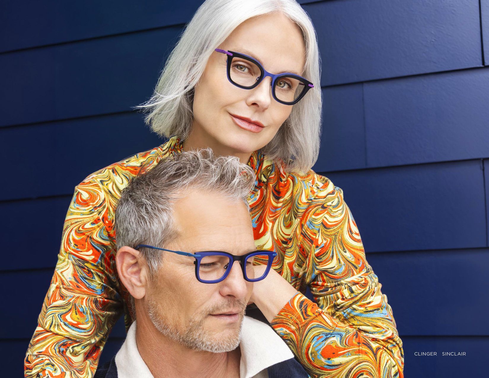 Two people with blue glasses posing against a dark blue wall, one standing behind the other.