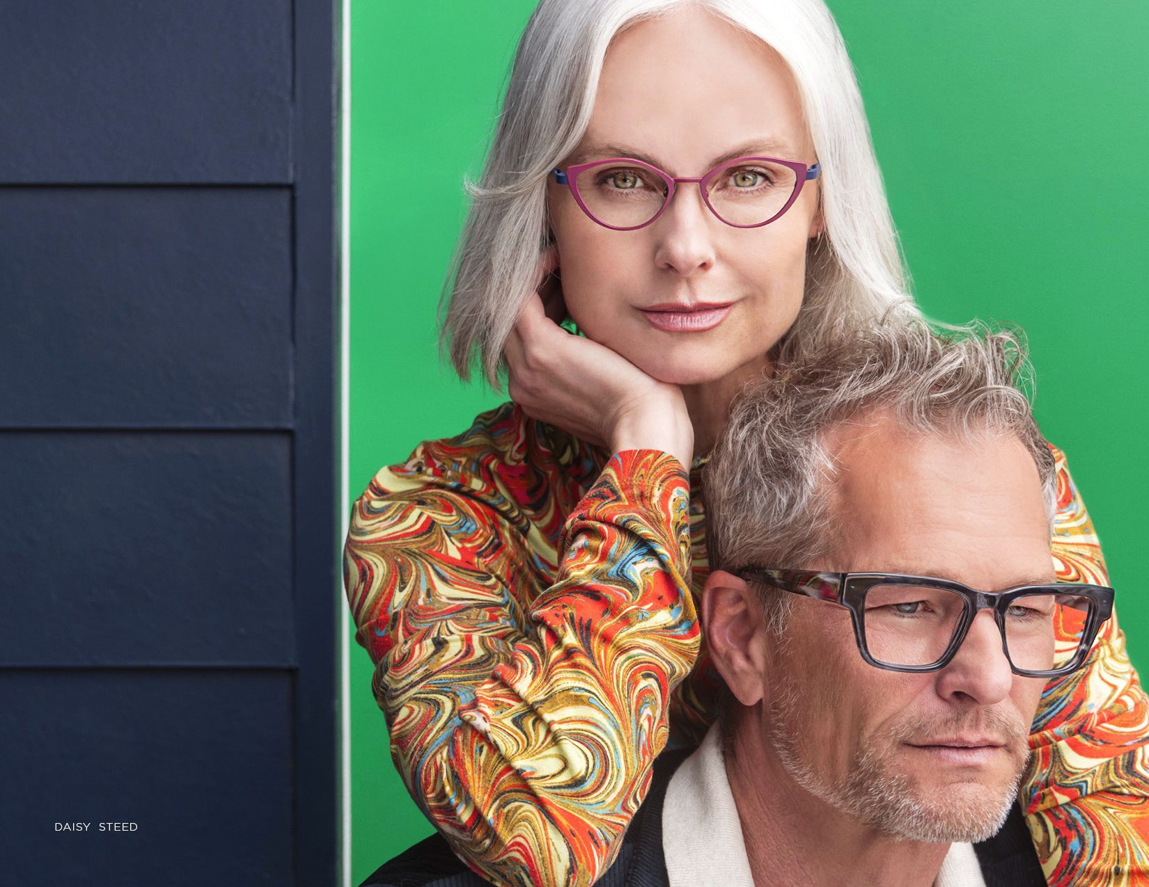 Two people with glasses posing against a green backdrop, one resting their head on the other’s shoulder.