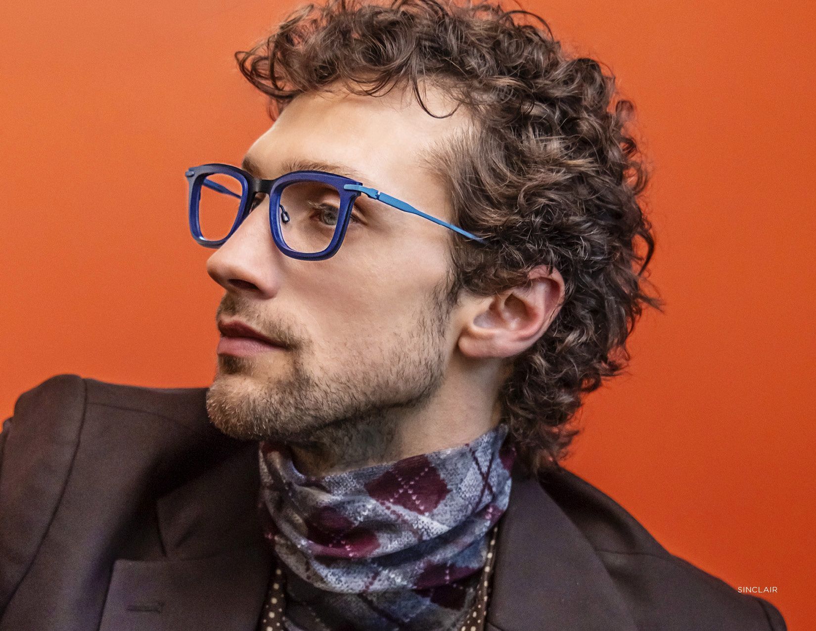 Man with curly hair and blue glasses against an orange background, looking to the side.