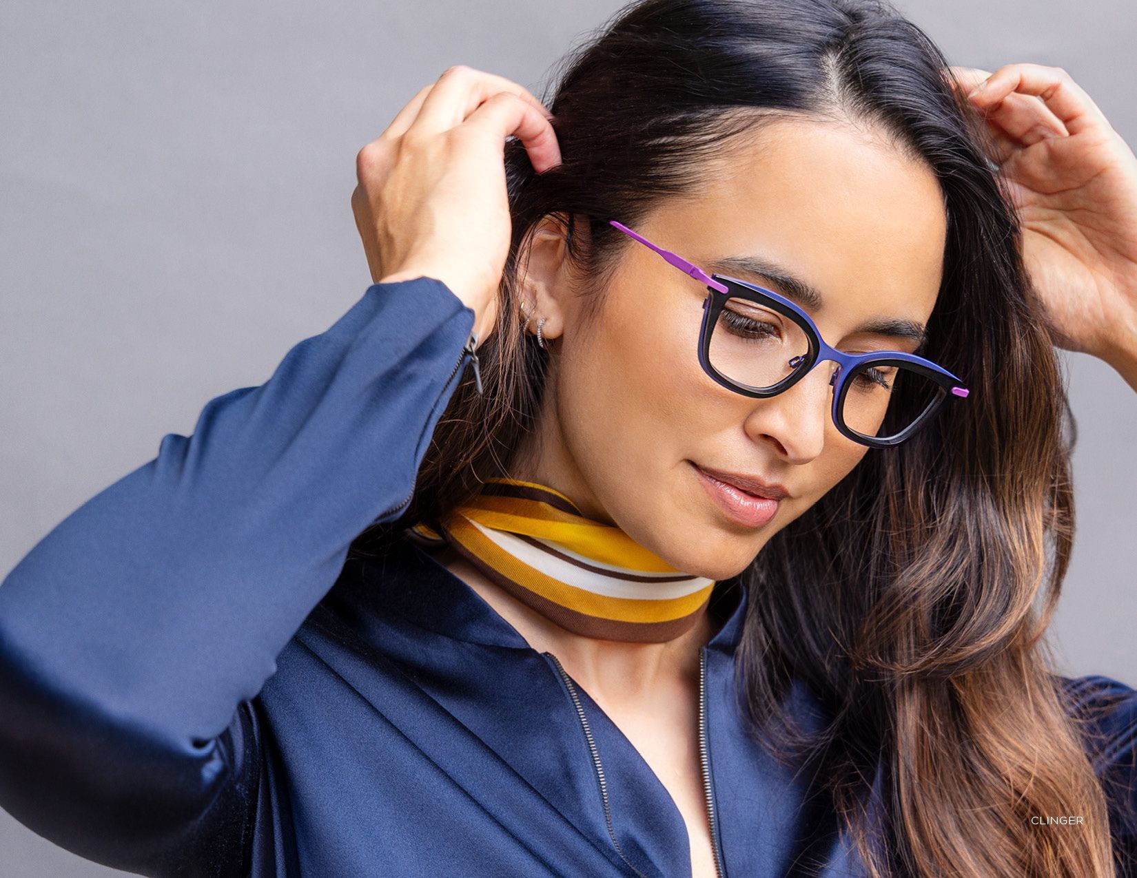 Woman adjusting her hair, wearing purple glasses and a striped scarf with a navy blouse.