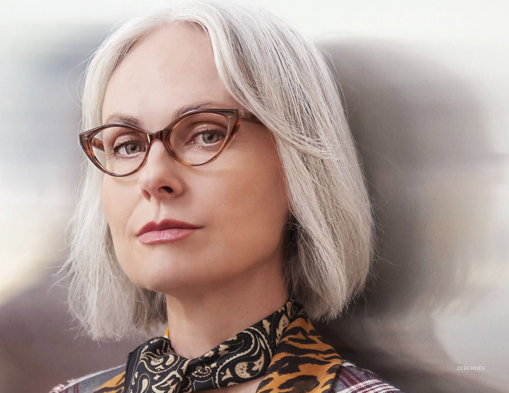 Portrait of a woman with white hair and glasses, wearing a patterned collar against a soft gray background