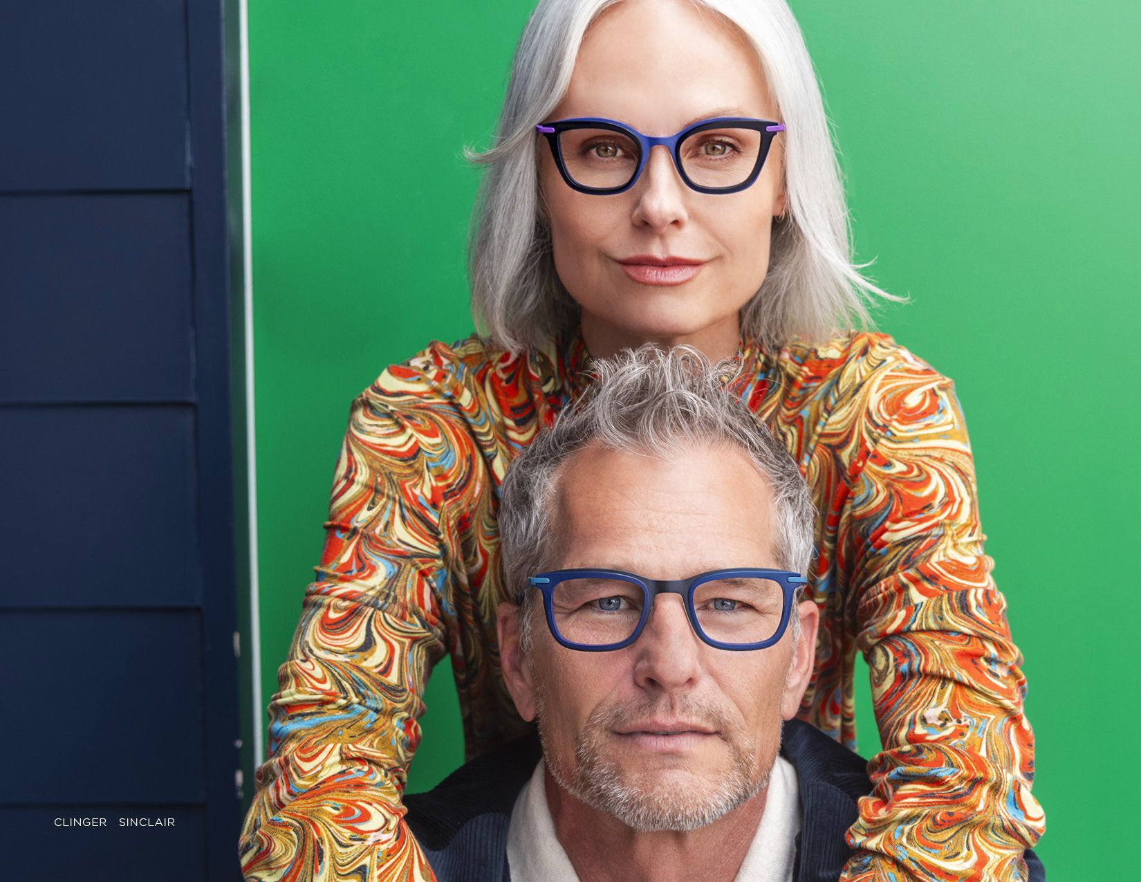 Two people wearing blue glasses pose closely against a green and dark blue background.