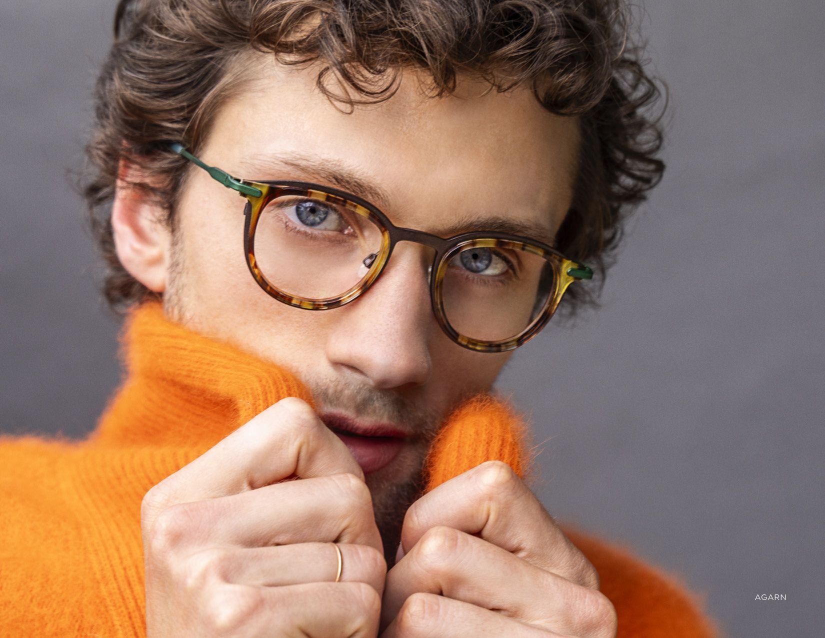 Close-up of a person in tortoiseshell glasses and an orange sweater, pulling the collar up against a gray background