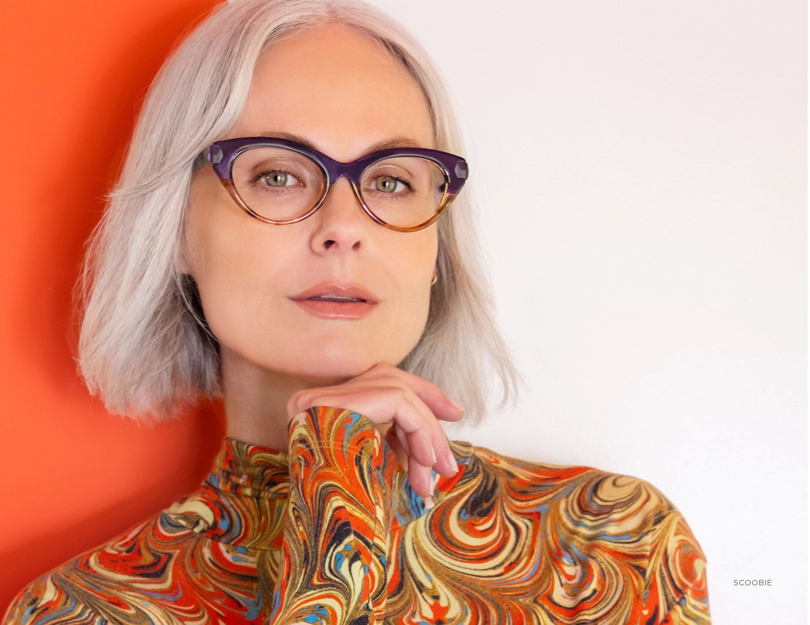 Portrait of a person with silver hair wearing patterned glasses and a colorful swirled turtleneck against a coral background