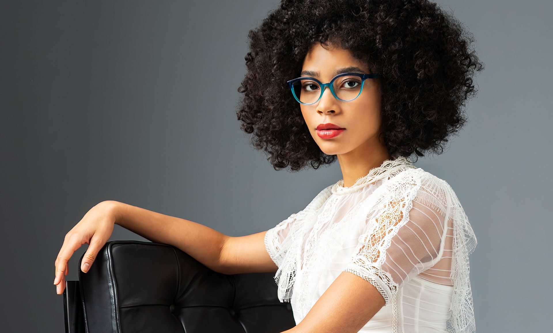 A woman wearing glasses and a white dress is sitting on a chair.