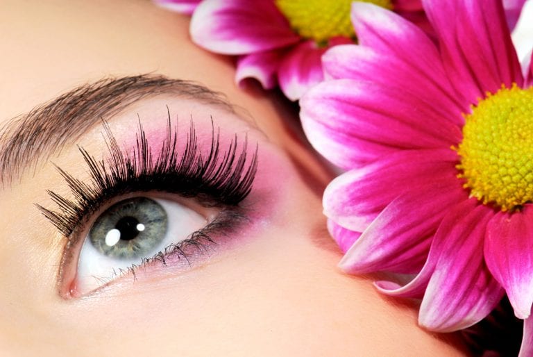 A close up of a woman 's eye with long eyelashes and a pink flower in the background.