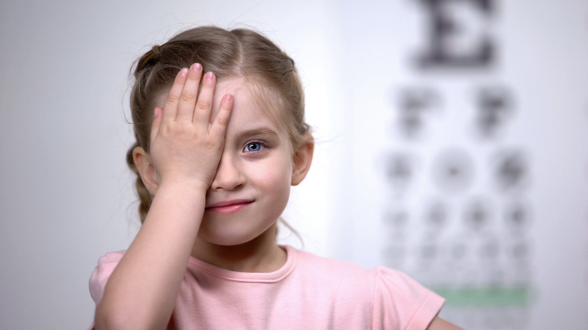 A little girl is covering her eye with her hand in front of an eye test chart.