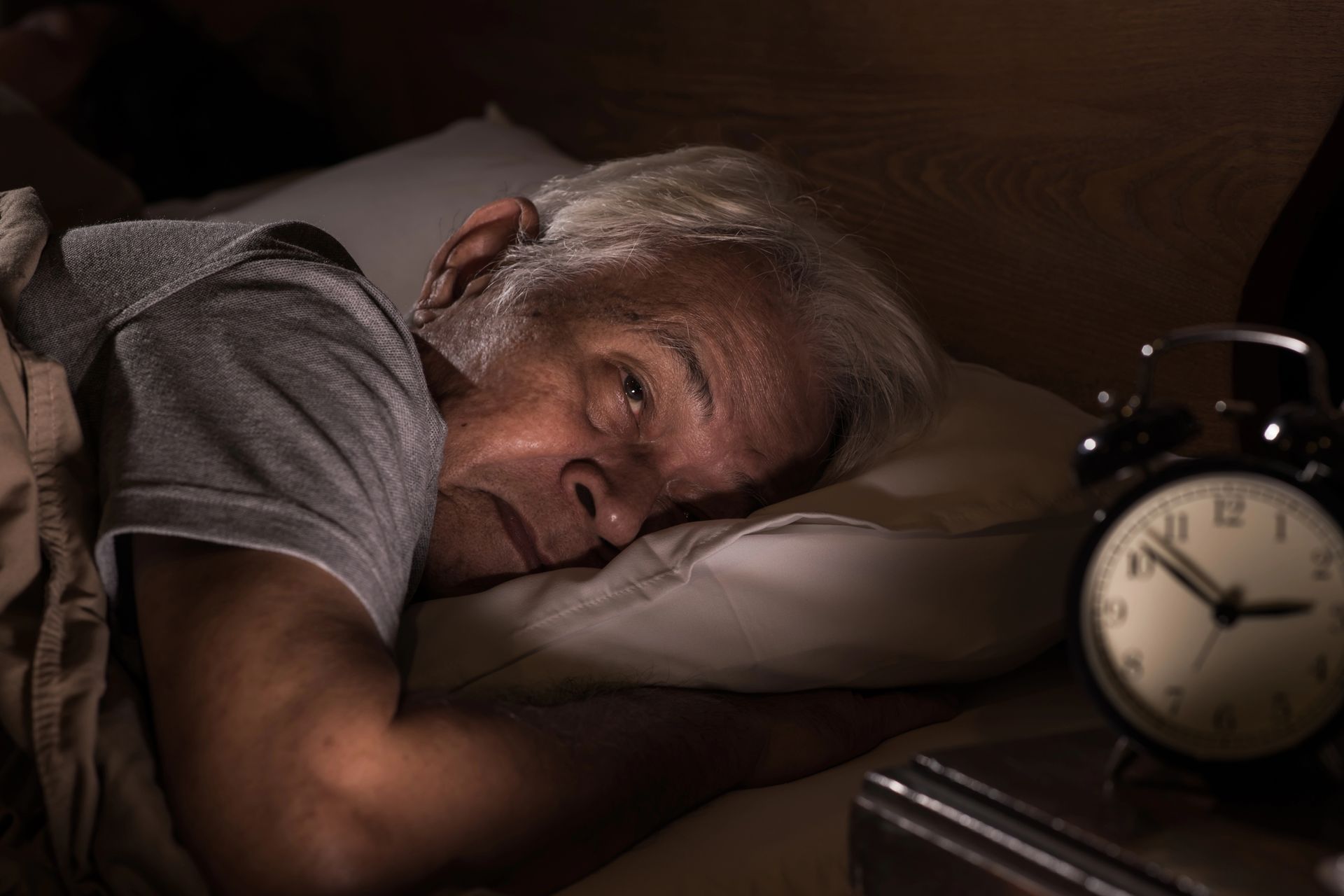 An elderly man is laying in bed next to an alarm clock.