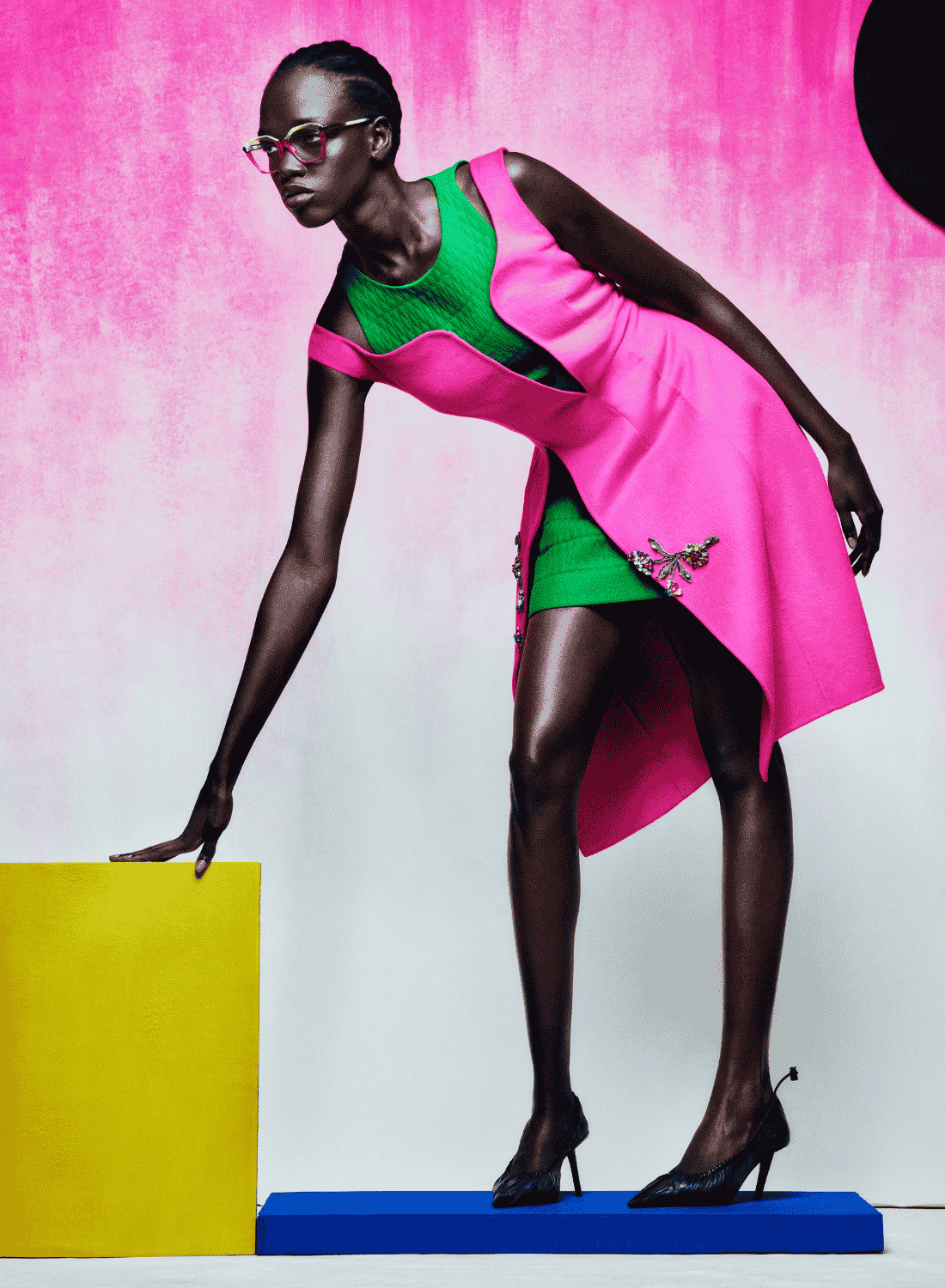 Fashion model in a bright pink and green outfit posing beside colorful blocks on a studio backdrop