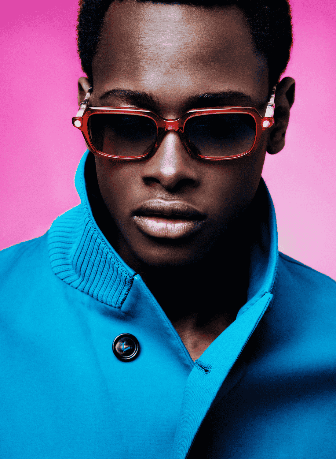 Portrait of a person in sunglasses and a bright blue coat against a pink background