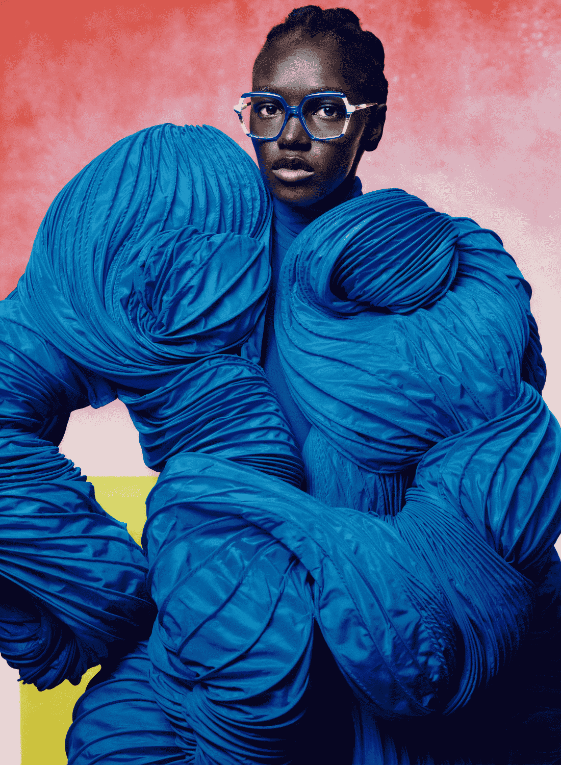 Person in a bright blue textured outfit against a pink background, wearing glasses, looking at the camera
